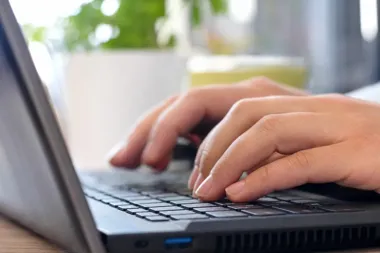 Dłonie osoby piszącej na klawiaturze laptopa na drewnianym stole w jasnym pomieszczeniu.